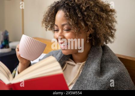 Donna sorridente avvolta in un libro di lettura scialle e in una tazza Foto Stock