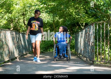 Adolescente (16-17) in sedia a rotelle con un amico che cammina nel parco Foto Stock