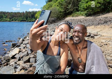 Coppia sorridente avvolta in asciugamani e scattata un selfie sulla riva del lago, Yorkshire, Regno Unito Foto Stock