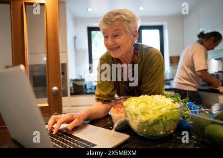 Donna anziana sorridente che guarda un notebook in cucina Foto Stock