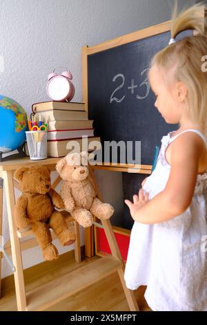 bambina, bionda che gioca a scuola, la piccola maestra insegna lezione, casa-ufficio studentesca, sveglia, libri, orsacchiotto, gesso, globo, concetto di ed Foto Stock