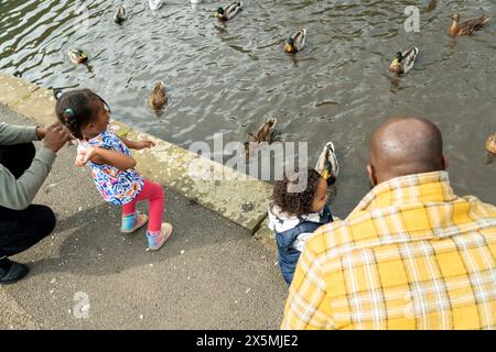 Due uomini con figlie che danno da mangiare alle anatre sullo stagno nel parco Foto Stock