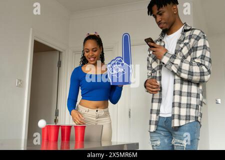 Donna che tira e uomo che usa lo smartphone mentre gioca a beer pong a una festa a casa Foto Stock