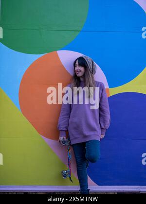 Ritratto di donna in piedi con lo skateboard contro una parete multicolore Foto Stock