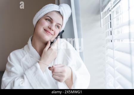 Donna sorridente in accappatoio e asciugamano turbante che parla sullo smartphone Foto Stock