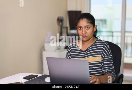 Una giovane donna indiana, vestita con un abbigliamento business casual, siede a una scrivania nel suo ufficio domestico e si concentra sul suo lavoro su un computer portatile Foto Stock