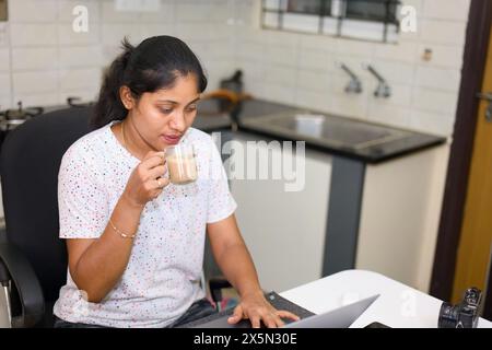Una giovane donna indiana felice si gode una pausa caffè mentre lavora in modo produttivo sul suo computer portatile in una cucina luminosa trasformata in un ufficio domestico. Freelancin Foto Stock