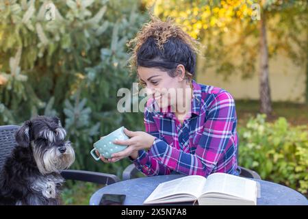 Bella giovane donna seduta in giardino con un cane sulla sedia, leggendo un libro e bevendo tè Foto Stock