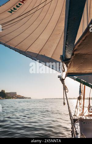 Una vista da una feluca sul Nilo vicino a Luxor mostra la vela che cattura la luce del pomeriggio, con acque calme che si estendono verso la lontana riva del fiume. Foto Stock