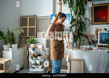 Allegra pittrice che lega il grembiule per prepararsi al lavoro creativo. Artista femminile al laboratorio d'arte Foto Stock