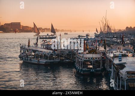 Dozzine di barche decorate sono attraccate lungo il Nilo a Luxor al tramonto, con feluche che navigano in lontananza sotto un cielo arancione e rosa. Foto Stock