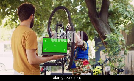 Giovane uomo con laptop che mostra schermo verde mentre la signora afroamericana attacca la ruota al telaio della bicicletta nel cortile. Personal computer con modello cromakey isolato tenuto da maschio caucasico. Foto Stock