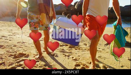 Immagine di palloncini a forma di cuore che si muovono su amici maschi caucasici che trasportano refrigeranti in spiaggia Foto Stock