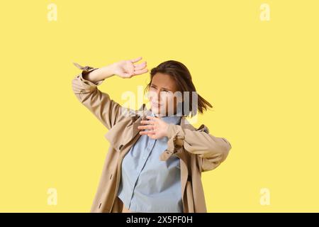 Giovane donna in cappotto con vento forte su sfondo giallo Foto Stock