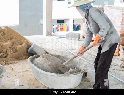 Operaio che utilizza il retroescavatore per mescolare manualmente sabbia e cemento nel cantiere Foto Stock