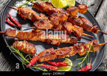 delizioso kebab di maiale succoso su piatto grigio con rosmarino, lime e peperoncino rosso secco su un tavolo rustico in legno, primo piano Foto Stock