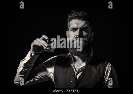 L'uomo con la barba regge il bicchiere con alcol. Barman o degustatore dal volto curioso regge un bicchiere di cognac. Uomo barbuto che indossa un vestito e beve whisky Foto Stock