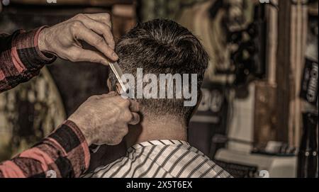 Primo piano di un barbiere professionista che lavora dando un taglio di capelli ad un uomo. Il cliente hipster si fa tagliare i capelli. Mani di barbiere con regolacapelli, primo piano Foto Stock