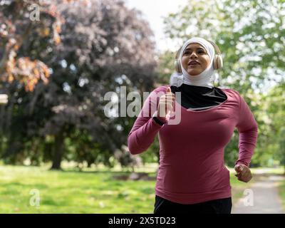 Regno Unito, Sutton, donna con foulard e cuffie che fa jogging nel parco Foto Stock