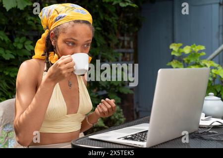 Giovane donna che beve il tè mentre lavora su un computer portatile in giardino Foto Stock