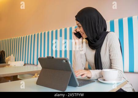 Giovane donna in hijab che lavora su un tablet e parla al telefono in un bar Foto Stock