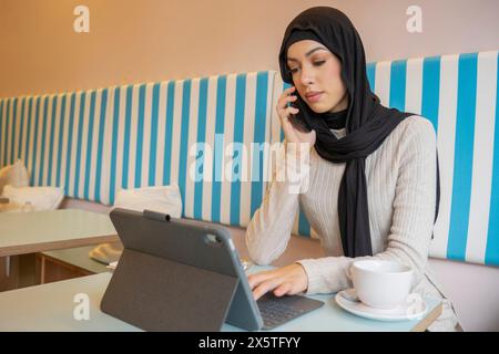Giovane donna in hijab che lavora su un tablet e parla al telefono in un bar Foto Stock