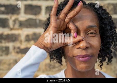 Ritratto di donna che guarda attraverso il segno OK fatto con le dita contro il muro di mattoni Foto Stock