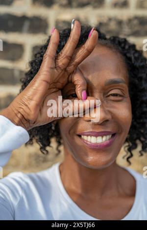 Ritratto di donna che guarda attraverso il segno OK fatto con le dita contro il muro di mattoni Foto Stock