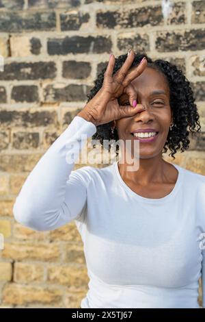 Ritratto di donna che guarda attraverso il segno OK fatto con le dita contro il muro di mattoni Foto Stock