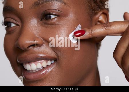 Primo piano di una donna sorridente che applica la crema per il viso sulla guancia Foto Stock