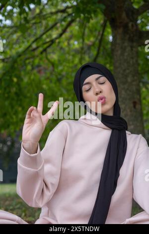 Ritratto di donna in hijab che fa volti e mostra un segno di pace nel parco Foto Stock
