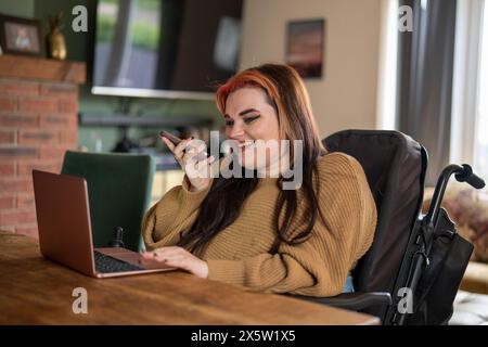 Donna in sedia a rotelle che parla al telefono e utilizza un computer portatile Foto Stock
