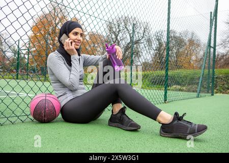 Donna in hijab con smartphone e bottiglia d'acqua seduta sul campo da basket Foto Stock