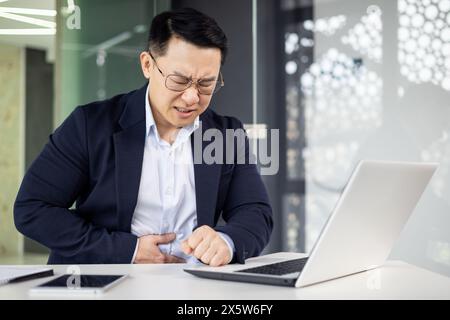 Un uomo d'affari asiatico di mezza età sente un improvviso dolore allo stomaco mentre si concentra sul lavoro su un notebook in un ambiente di lavoro elegante. Foto Stock
