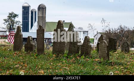 Immagine suggestiva che cattura le lapidi in un cimitero storico, sullo sfondo di una fattoria rurale con silos e fienili, sotto un cielo nuvoloso. Foto Stock