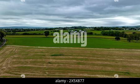 Questa vista aerea mostra un suggestivo paesaggio rurale sotto un cielo coperto, caratterizzato da zone contrastanti di lussureggianti campi verdi e aree di raccolta. Foto Stock
