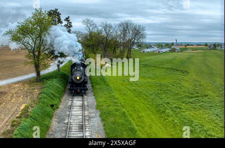 Colpo d'atmosfera di un treno a vapore d'epoca che attraversa un paesaggio rurale, emettendo fumo intenso, con uno sfondo di campi appena arati e vegetazione lussureggiante. Foto Stock