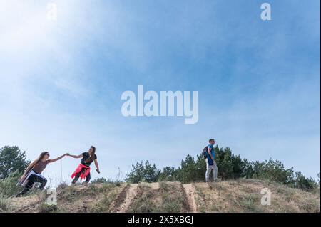 Una ragazza aiuta un'altra ragazza su una collina. Tre amici in un'escursione. Grandangolo. Contro il cielo. Foto Stock