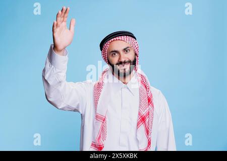 Uomo musulmano sorridente vestito con abiti tradizionali che solleva il ritratto di uno studio a mano. Persona islamica che saluta un amico, sventolando con il braccio e guardando la macchina fotografica con un'espressione allegra Foto Stock