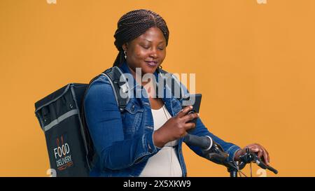 Donna nera attiva che consegna cibo in bicicletta con il cellulare in mano. Donna afroamericana che controlla l'app di consegna su smartphone per il trasporto del pacco. Scatto di ritratti, sorridendo alla fotocamera. Foto Stock