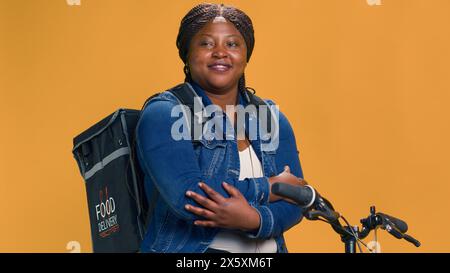 Giovane ciclista donna seduta in bicicletta con la borsa per la consegna del cibo sulla schiena in attesa di consegnare i piatti da asporto. Messaggero di bici afroamericano riposa prima di inviare i pacchi. Scatto a mano libera. Foto Stock