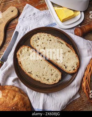 Fette di pane fresco a base di lievito madre su un piatto con burro su un tavolo di legno Foto Stock