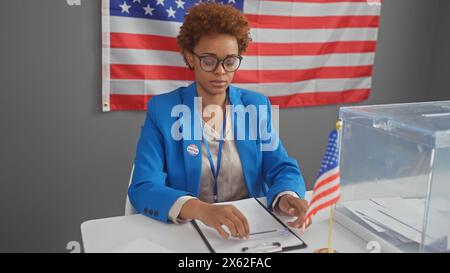 Una donna afroamericana che indossa un distintivo siede pensiamente in una postazione elettorale, con la bandiera degli Stati Uniti sullo sfondo. Foto Stock