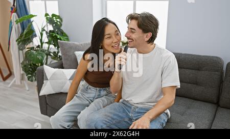 Una felice coppia interrazziale in abbigliamento casual canta all'interno, utilizzando un telecomando tv come microfono improvvisato, mostrando affetto e intrattenimento a hom Foto Stock