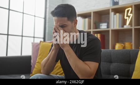 Un giovane angosciato ispanico si copre il viso con le mani, seduto in un moderno soggiorno di un appartamento. Foto Stock