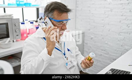 Un anziano con un camice bianco da laboratorio esamina i farmaci mentre parla al telefono in un laboratorio. Foto Stock