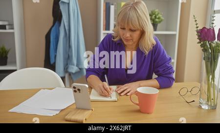 Una donna bionda scrive in un taccuino a casa, circondato da fiori, una tazza e bicchieri su un tavolo di legno. Foto Stock