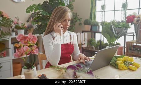 Una donna bionda fiorista in grembiule rosso che utilizza un computer portatile e un telefono in mezzo a fiori vivaci all'interno di un negozio di fioristi. Foto Stock