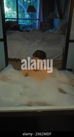 Una giovane donna ispanica si rilassa in un bagno di bolle con la camera da letto di un hotel di lusso sullo sfondo. Foto Stock