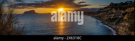 Il tramonto dorato dipinge il cielo mentre si tuffa sotto l'orizzonte montuoso, riflettendo sulle acque del Mediterraneo vicino alle scogliere costiere di Calpe, Spagna Foto Stock